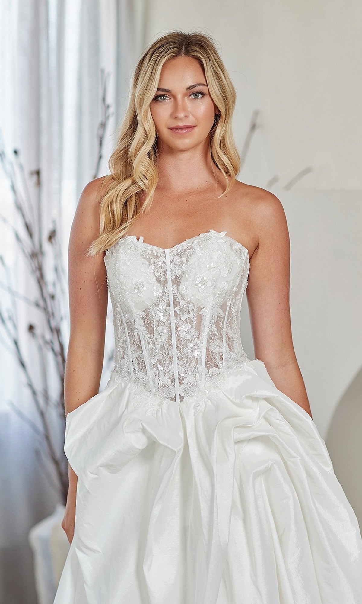 A woman stands indoors wearing the Amelia Couture 2087, a long prom dress with a strapless lace corset bodice and voluminous satin A-line skirt.